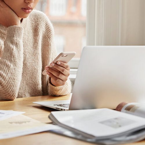 Serious female worker sits at desktop with laptop computer, surrounded with papers and journals, types sms message on smart phone device, connected to wireless internet, poses near window indoor